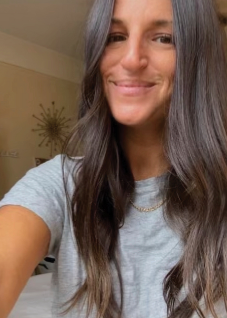 A person with long, dark hair smiling in a room with modern decor on the wall.