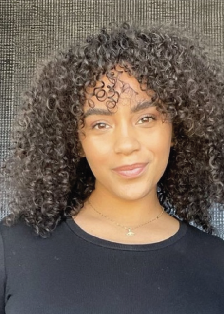 Person with curly hair smiling, wearing a black top and necklace, against a textured dark background.